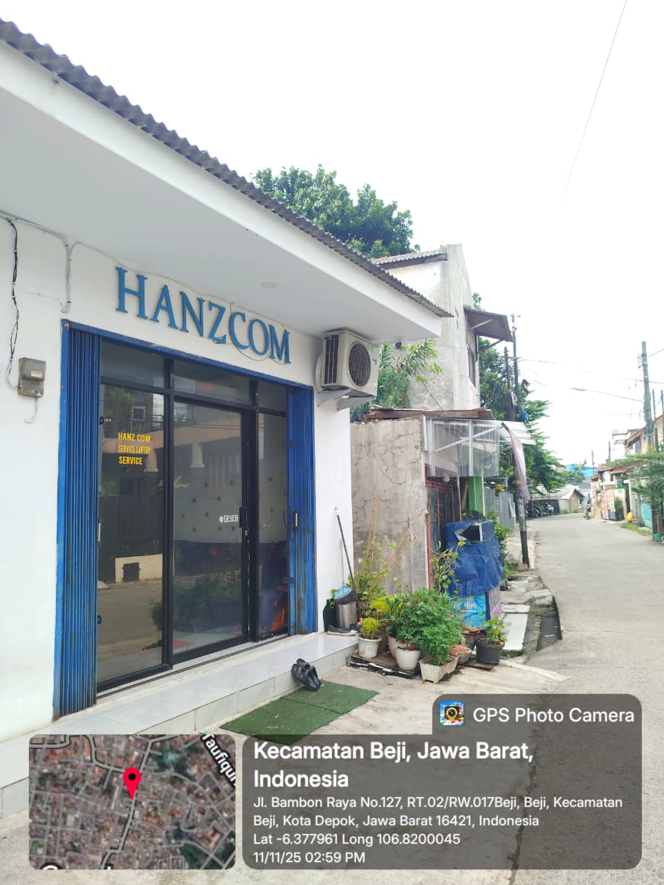 Tampak bangunan Hanz Com Laptop di Beji Depok dengan fasad biru, pintu kaca geser, pot tanaman, dan suasana gang sekitar di Jl. Bambon Raya.”